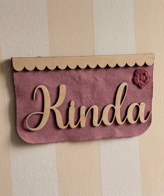 Personalized dusty rose textured fabric name banner with natural wooden lettering, a scalloped wooden top trim, and a decorative crochet flower, perfect for a nursery or children’s bedroom.