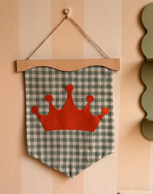 Little wavy fabric wall banner in sage green gingham with a red felt crown appliqué, displayed on a wooden hanger in a kids’ room.