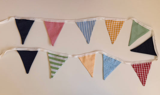 Multicolor fabric bunting garland featuring navy, green, blue, red and mustard flags in gingham, striped and patterned fabrics, finished with white binding for kids’ room decor.