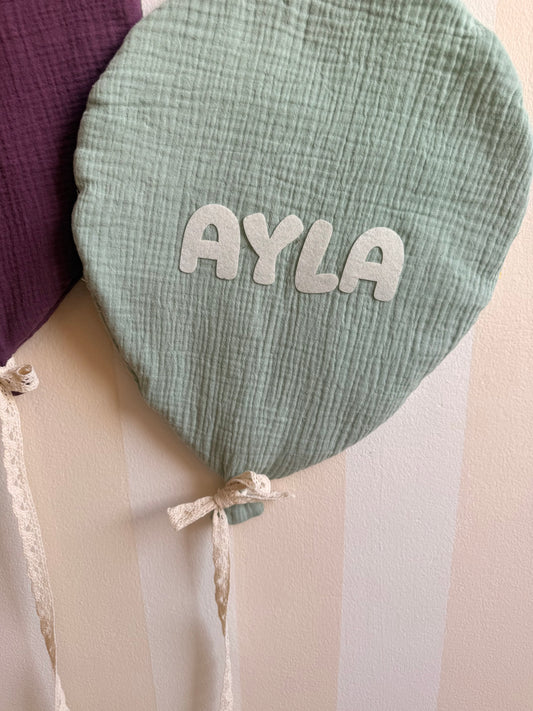 Close-up of a sage green balloon-shaped cushion with the personalized name Ayla in white lettering, made from textured fabric with a decorative lace ribbon.