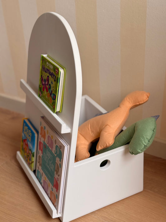 White wooden kids’ book and toy organizer with a rounded handle, front-facing book display shelves, and storage space for colorful dinosaur cushions.
