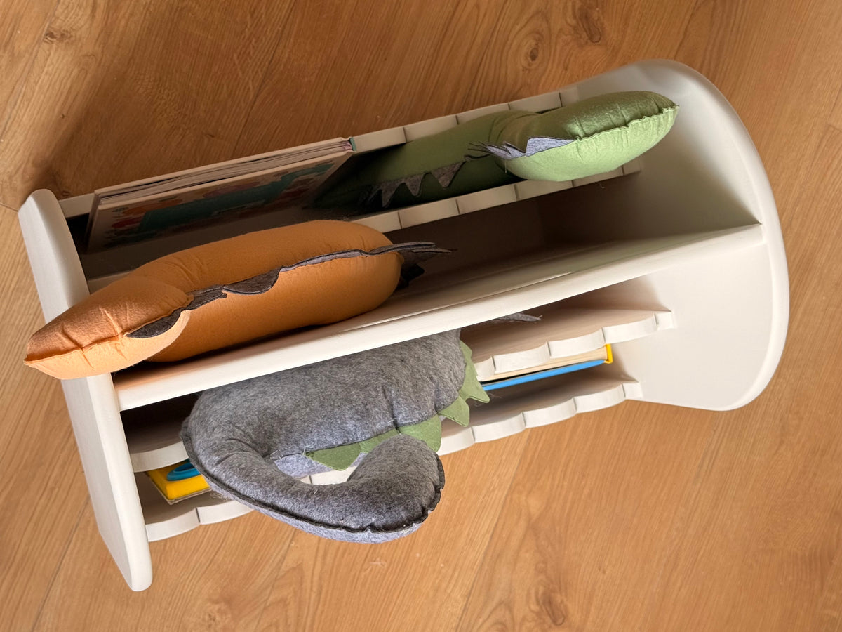 Top view of a white wooden kids’ book organizer with multiple compartments holding children’s books and green, grey, and orange dinosaur cushions.