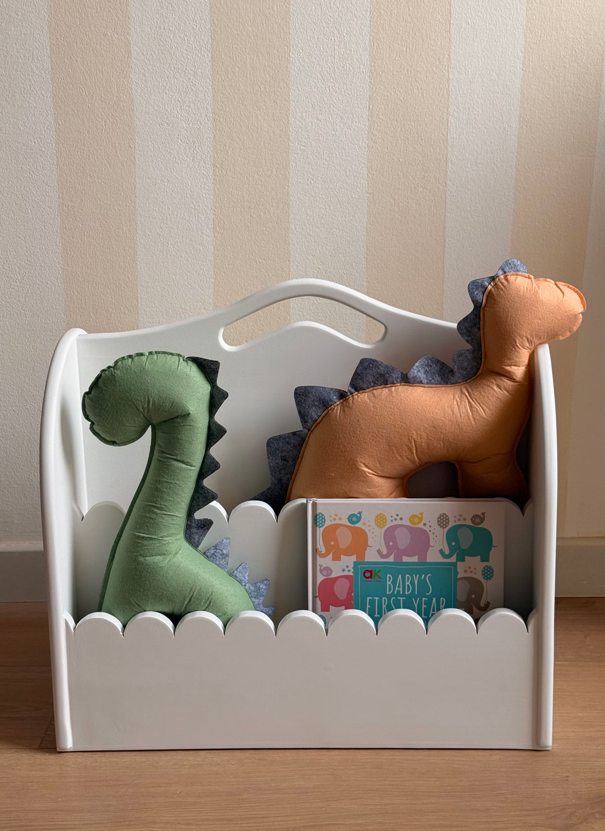 White wooden kids’ book organizer with scalloped front edges, filled with colorful children’s books and handmade green and orange dinosaur cushions.
