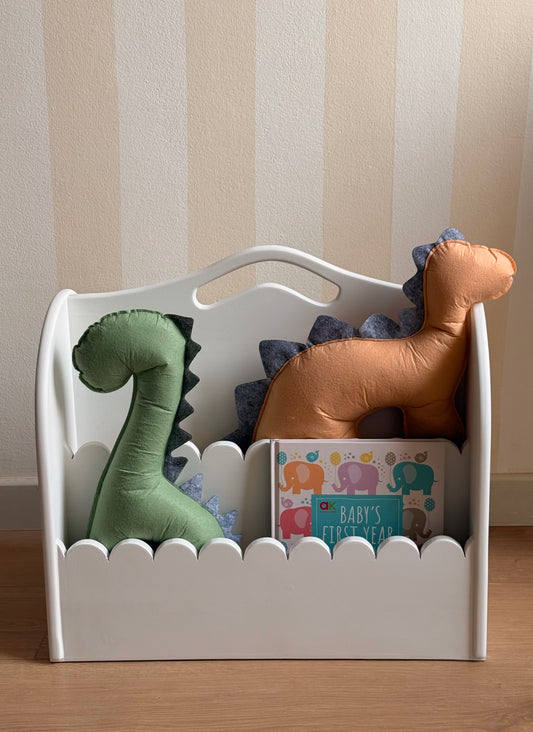 White wooden kids’ book organizer with scalloped front edges, filled with colorful children’s books and handmade green and orange dinosaur cushions.