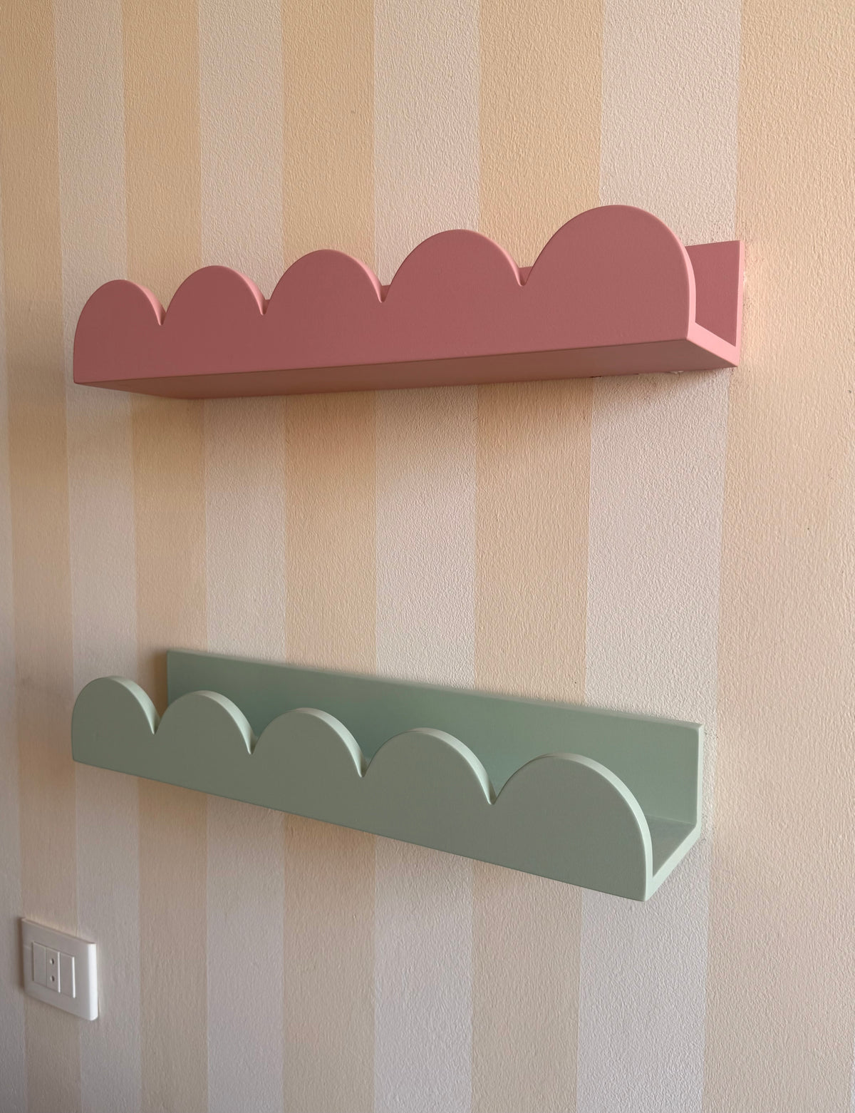 Two cloud-shaped wall shelves mounted on a beige and cream vertically striped wall. The top shelf is dusty pink, and the bottom shelf is sage green.