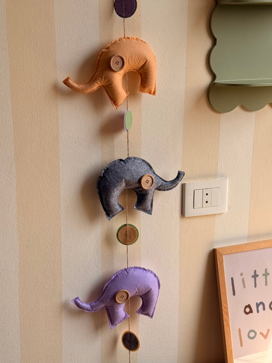Close-up of a handmade felt elephant garland with orange, charcoal grey, and lavender elephants, wooden button embellishments, and multicolored felt circles strung vertically.