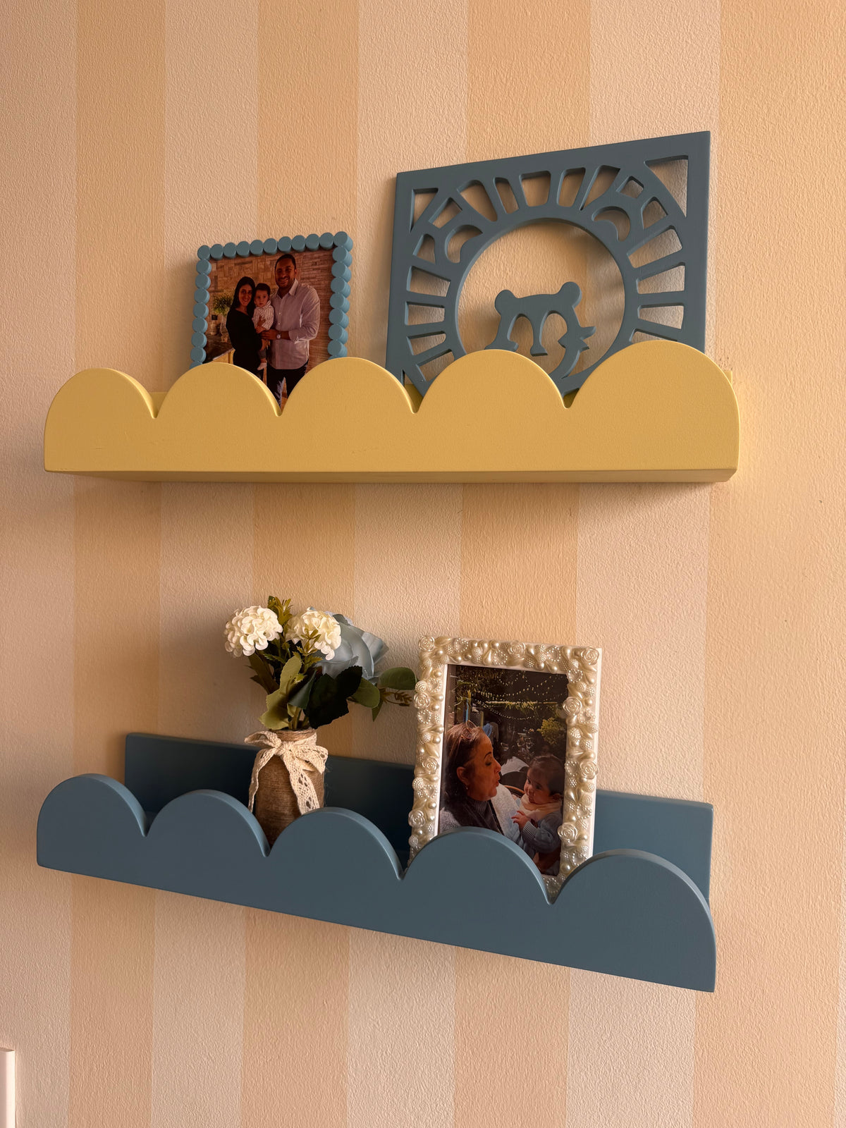 A two-tier cloud-shaped wall shelf set in yellow and slate blue, styled with picture frames and a vase of flowers against a beige and white striped wallpaper background.