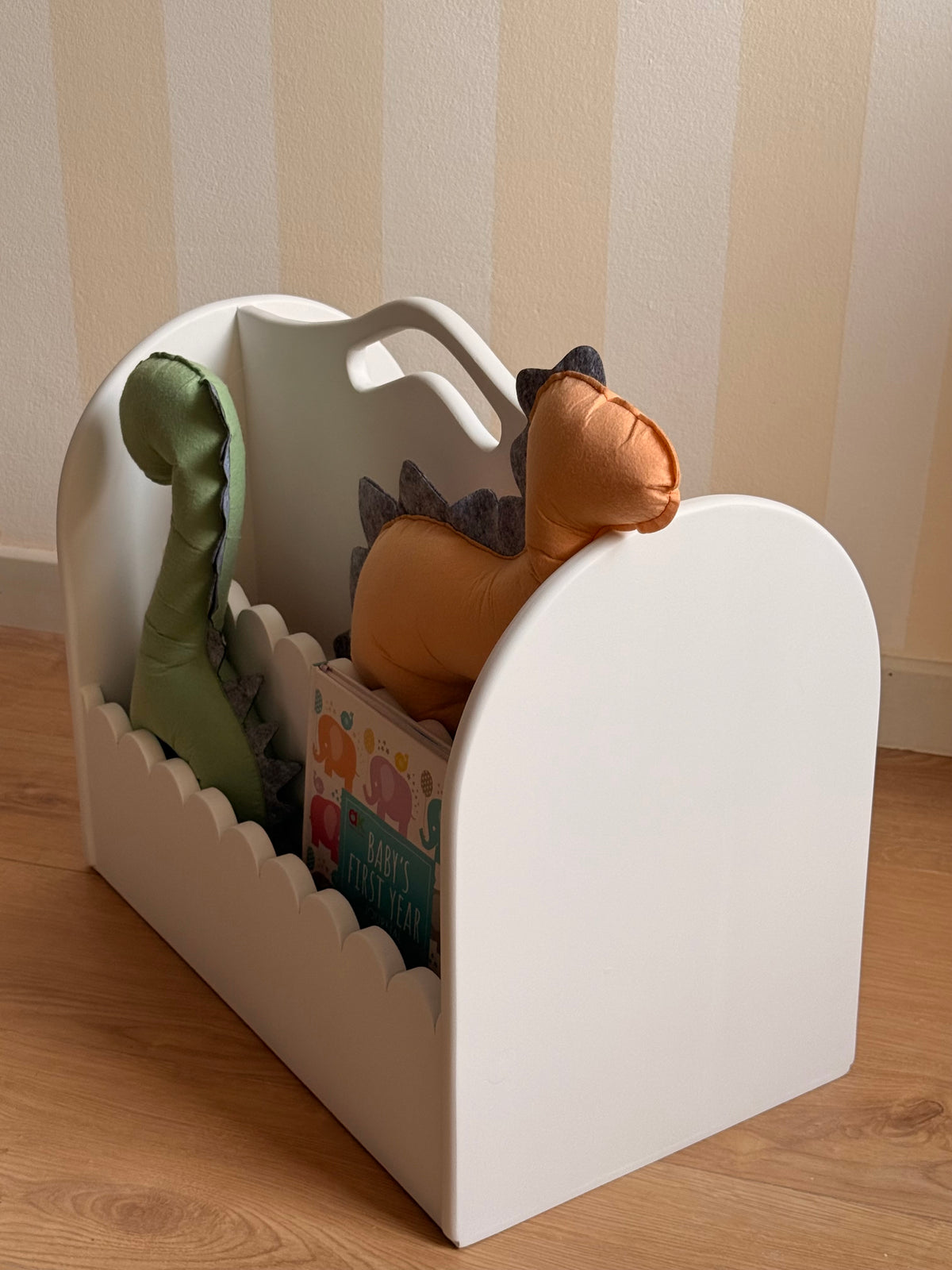 Side view of a white wooden children’s book organizer with rounded edges and a carry handle, displaying kids’ books and soft dinosaur cushions in a nursery.
