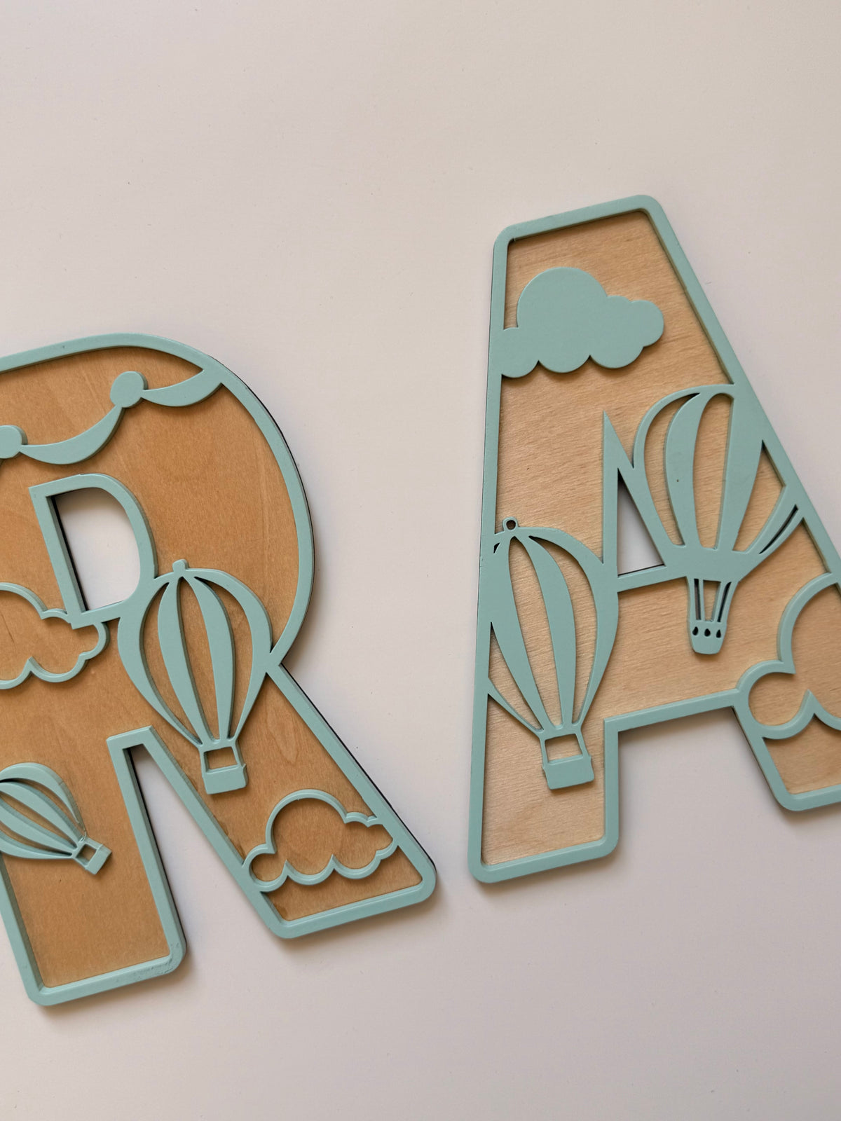 Close-up of personalized wooden name letters with mint green outlines, natural wood backgrounds, and detailed hot air balloon and cloud designs for a children’s room.