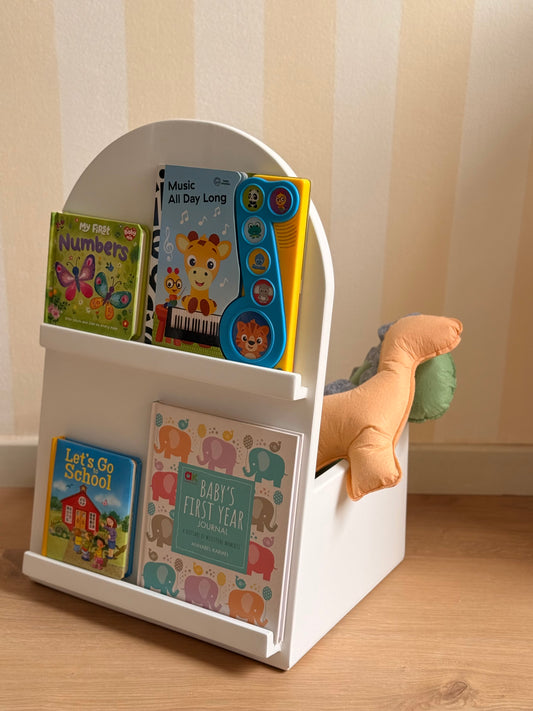 White wooden children’s book organizer with two front-facing display shelves, a rounded carry handle, and colorful children’s books, styled with a soft orange dinosaur cushion.