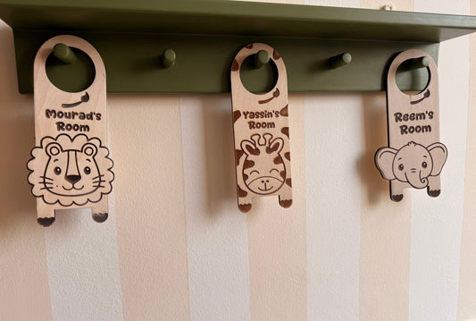 Olive green wooden wall rack with hooks displaying personalized wooden animal door hangers featuring lion, giraffe, and elephant designs with children’s room names.
