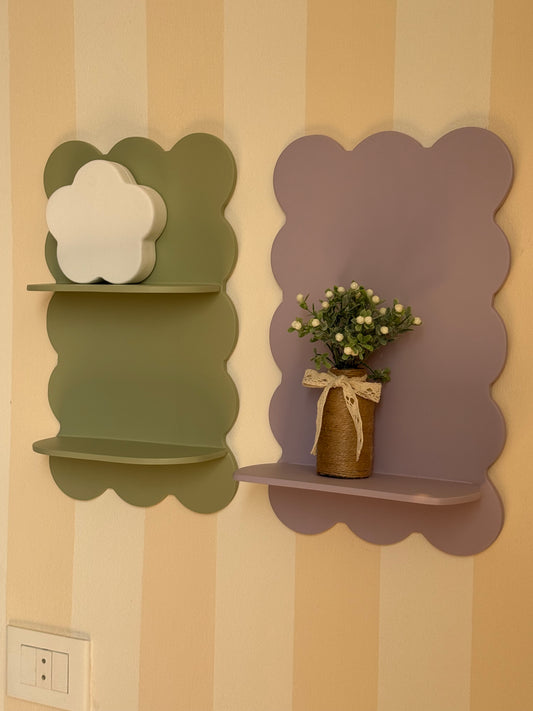 Two scalloped cloud-shaped wall shelves mounted on a cream and tan striped wall. The left shelf is olive green with a white cloud-shaped decor piece, and the right shelf is dusty lavender with a small potted plant in a brown vase.