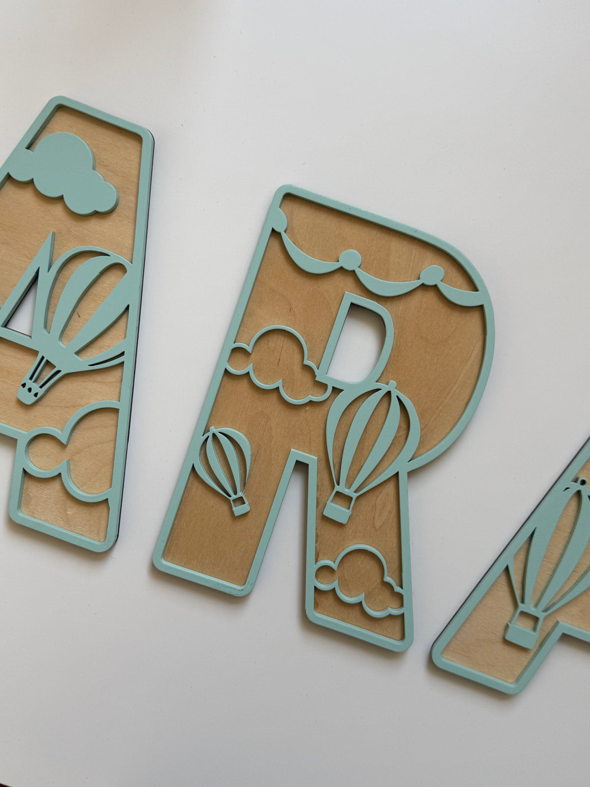 Close-up of a mint green and natural wood personalized letter featuring decorative hot air balloons and clouds, part of a custom wooden name sign for a nursery.