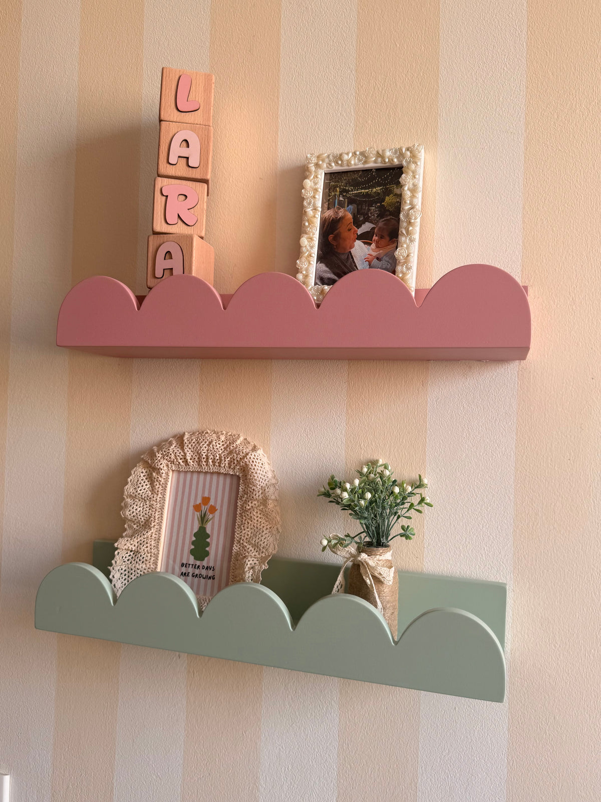 Two scalloped cloud-style wall shelves mounted on a cream and tan striped wall. The upper shelf is dusty pink and holds pink letter blocks spelling 'LARA' and a small framed photo.