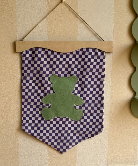 Little wavy fabric wall banner in purple gingham with a green felt teddy bear appliqué, displayed on a wooden hanger in a kids’ room.